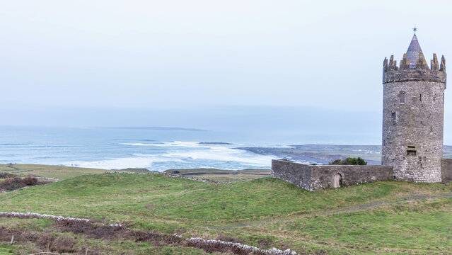 Aerial view of Doonagore Castle, a historic stone tower, situated on a grassy hill overlooking the Atlantic Ocean with waves crashing on the shore in Doolin, County Clare, Ireland.