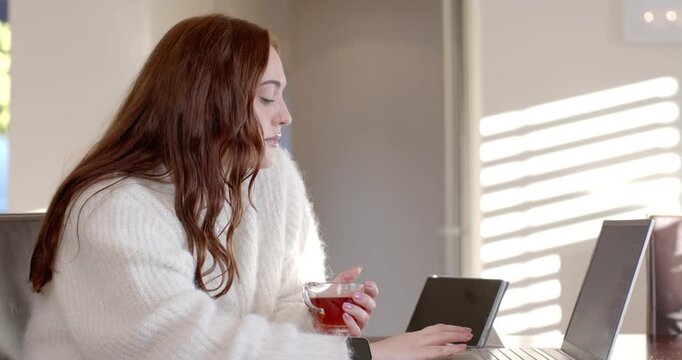 Woman in sweater working at sunlit table, reading tablet and tapping laptop, holding cup