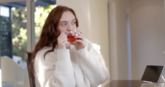 Woman in 20s pausing laptop at home, sipping tea from glass cup by window, wearing cardigan