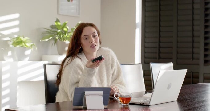 Adult woman raising phone to mouth, sending at home table in sweater checking tablet and laptop