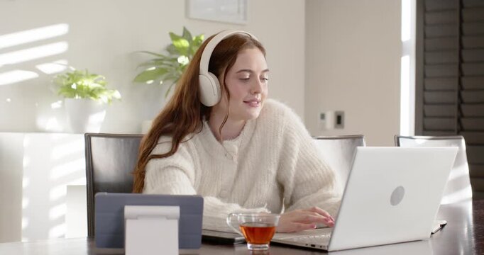 Adult woman wearing white headphones and cream sweater typing and speaking to laptop, tea visible