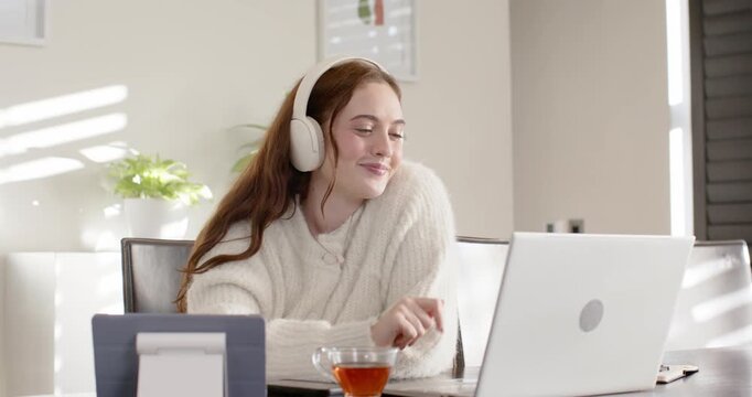 Woman in cream sweater greeting laptop after call alert, adjusting headset joining meeting at home