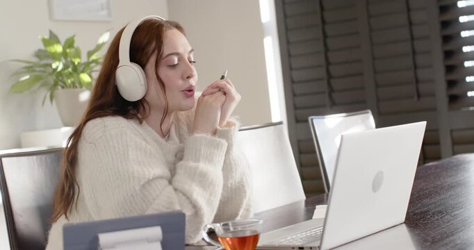 Woman sitting at dining table prompted by laptop wearing headphones holding pen practicing speech