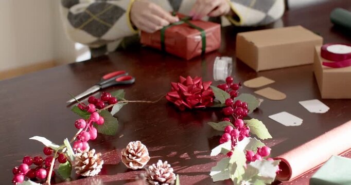 Hands entering frame picking up red-wrapped box, woman in 20s tying ribbon preparing holiday gift