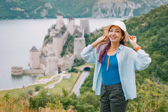 Adventurous traveler adjusts her hat at a hilltop viewpoint, Golubac Fortress looming behind
