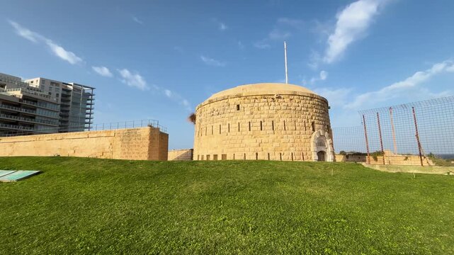 circular limestone fort tower crowns grassy embankment chain link fence high rise skyline behind flagpole clear sky handheld footage malta coastal fortification historic gun battery hilltop viewpoint