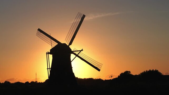 Silhouette of a traditional Dutch windmill with blades turning against a beautiful orange sunset sky in the Netherlands.