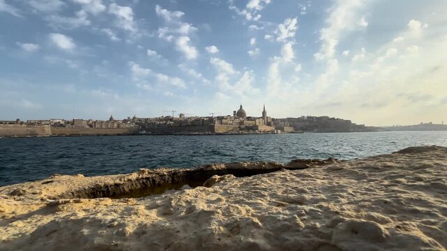 waves crash against golden limestone rocks valletta skyline rises across water domes spires fortress walls fort st elmo handheld footage coastal malta capital seascape rugged shoreline turquoise sea