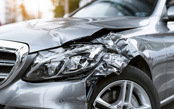 Close up view of damaged silver car after accident. Crumpled metal hood, smashed fender show heavy impact. Broken headlight, dented bumper near wheel at roadside. Vehicle needs expensive repair due