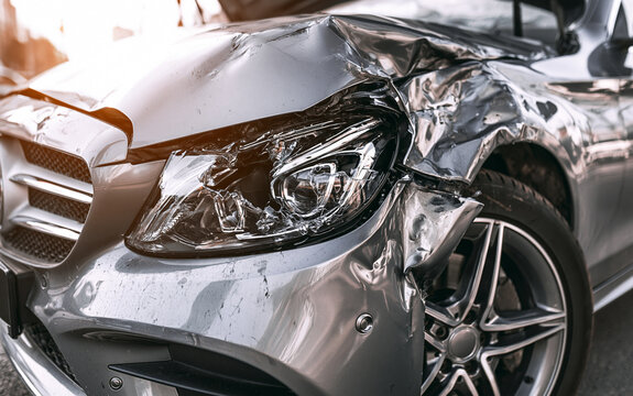 Close up view of damaged silver car after accident. Crumpled metal hood, smashed fender show heavy impact. Broken headlight, dented bumper near wheel at roadside. Vehicle needs expensive repair due
