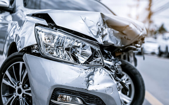 Close up view of damaged silver car after accident. Crumpled metal hood, smashed fender show heavy impact. Broken headlight, dented bumper near wheel at roadside. Vehicle needs expensive repair due