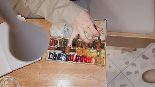 Close up of person hand selecting watercolor paints from open palette while reaching for sketchbook paper on wooden table. scene includes visible paint colors glass jar with liquid and soft