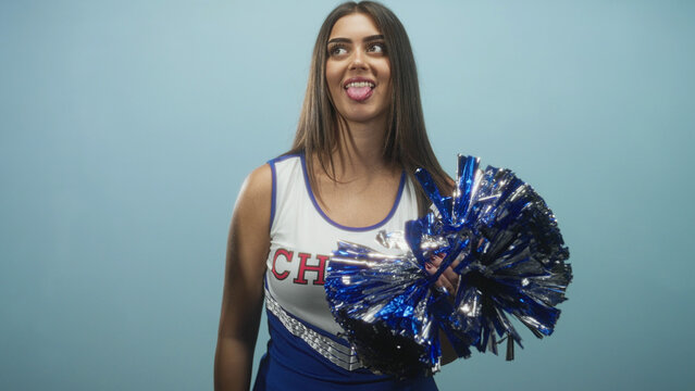 Cheerleader woman holding blue metallic pompom near chest and sticking tongue out in studio; playful team energy.