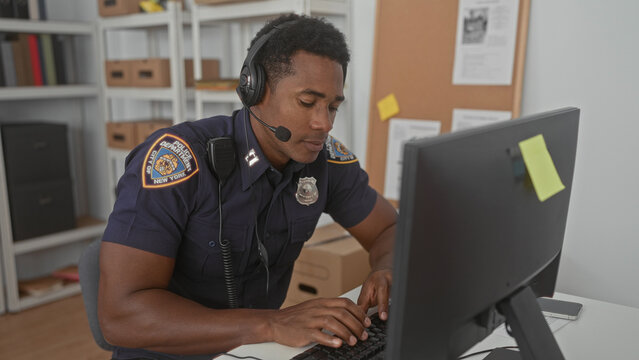 Man in police uniform wearing a headset typing on a keyboard at a building desk with monitor, badge, and cardboard boxes visible; focused duty.