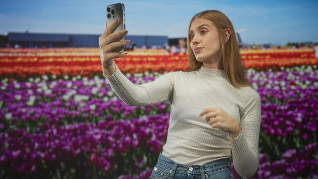 Woman holding smartphone for selfie with hand raised, posing in studio tulip field set; confidence travel nostalgia.