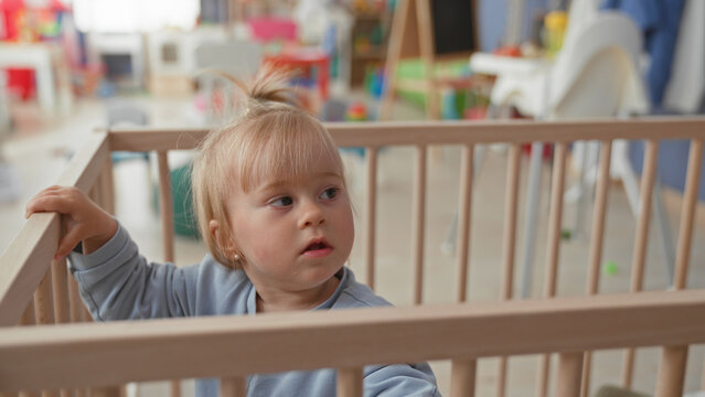 Toddler girl standing in crib holds rail and reaches up toward toy in nursery; curiosity play exploration.