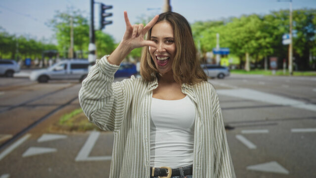Woman making i love you hand sign with bare hand and smiling at a city street intersection with cars and traffic light; joy travel.