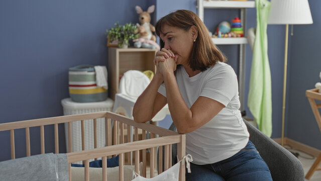 Woman clasping hands and leaning on wooden crib inside a baby room in a building, looking down at empty cradle and toys on shelf; quiet sorrow.