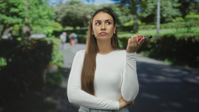 Young woman holding a vape with fingers near her mouth in a brief inhale gesture on a street path with blurred pedestrians and trees behind; contemplation calm.