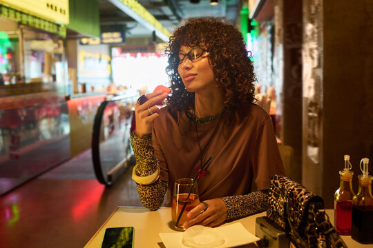 Young adult biracial woman with curly hair enjoying food in cafe with eyes closed. Image represents leisure and dining out, suitable for food blogs and lifestyle marketing