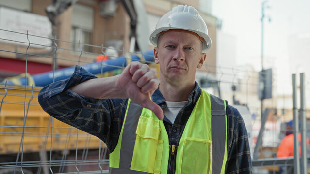 Man builder in hardhat and high visibility vest shows thumbs down with bare hand at construction building fence and equipment; safety concern disapproval.