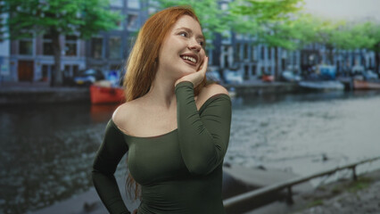 Fototapeta premium Redhead woman smiling, hand on cheek and bare shoulder by a street canal with moored boat and buildings; joyful carefree moment.