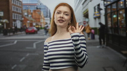 Fototapeta premium Redhead woman in striped off shoulder crop top making ok sign with right hand and bare shoulder visible on street; approval confidence.