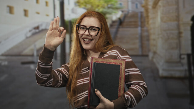 Redhead woman waving hand while holding a hardcover book on a stone street with stairs and arch; warm joy.
