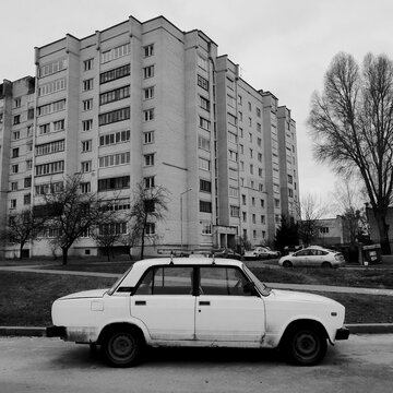 Black and white side view of a vintage classic white car parked on the street in front of a residential multi-story brick building