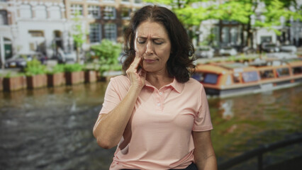 Fototapeta premium Woman touching cheek and wincing beside a canal boat on a street near buildings and trees; toothache pain concern.