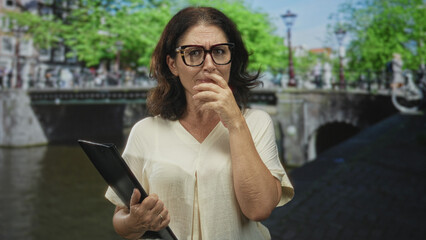 Middle aged hispanic woman holding a tablet case, hand to mouth and finger to lips for silence on a street  thoughtful reflection. © Krakenimages.com