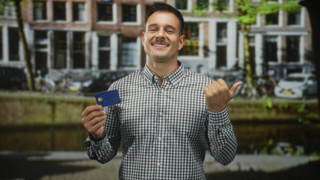 Hispanic man with moustache holding a creditcard and thumb pointing over shoulder, smiling with closed eyes in street by canal and building; confidence convenience.