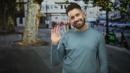 Man smiling and waving right hand, beard visible, wearing teal long sleeve shirt on street by trees, parked scooters and cars  friendly greeting. © Krakenimages.com