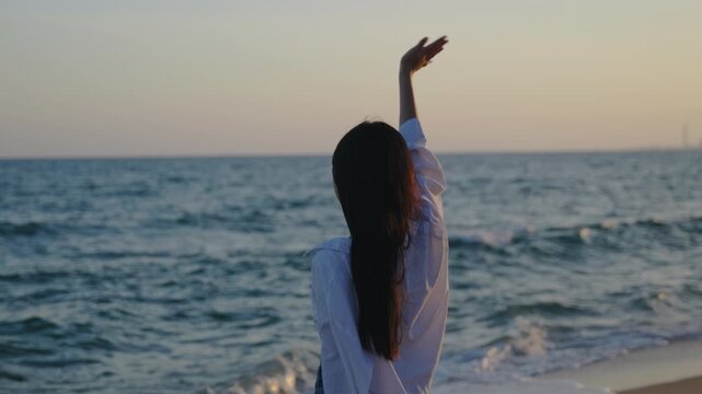 Happy young woman standing on beach waving hand in air during beautiful sunset, wearing casual white shirt while enjoying freedom and peace by ocean waves under warm evening sky.