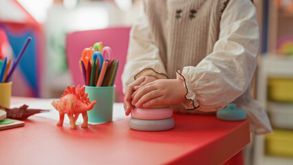Little blonde girl stacks pastel toy rings with bare hands on a preschool building table amid colored pencils and a dinosaur toy  playful curiosity. © Krakenimages.com