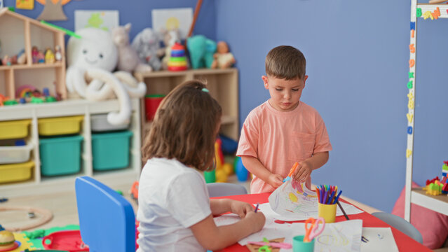 Boy holds orange scissors and girl draws on paper at a building playroom table with colored pencils and crafts; playful friendship.
