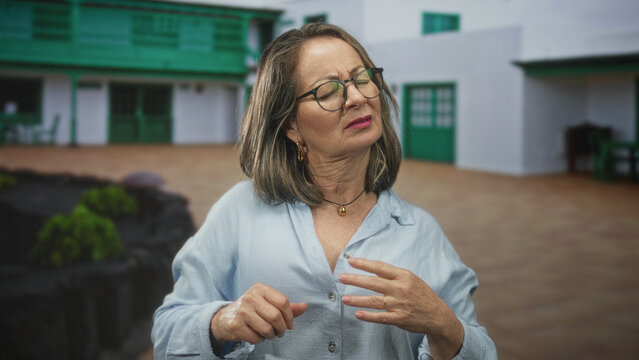 Woman with grey hair rubs shoulder and winces while standing in a building courtyard by green doors, wearing glasses and a light blue shirt; pain recovery.
