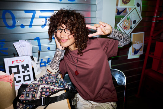 Portrait of young Asian woman with curly hair smiling and showing peace sign at cafe table. Image represents youth lifestyle and is suitable for modern dining app marketing