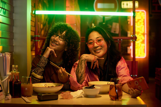 Portrait of young adult Caucasian woman and Asian woman smiling at table with Chinese food in neon lit cafe. Image represents youth lifestyle and is ideal for Asian restaurant marketing