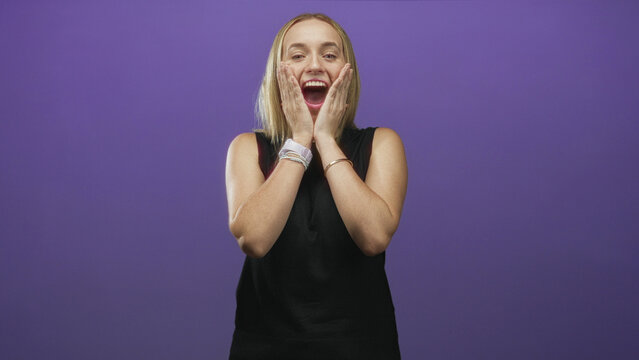 Woman with blonde hair, hands on cheeks and mouth open in surprised pose wearing sleeveless black top and bracelets against a purple studio wall; surprise joy.