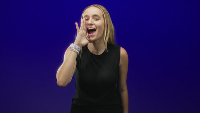 Woman cupping hand to mouth shouting in studio, blonde wearing black sleeveless top and smartwatch; enthusiasm announcement.