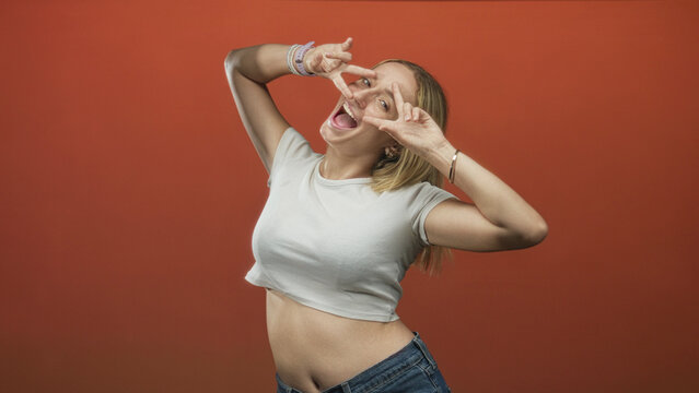 Woman exposing stomach wearing jeans and tshirt, making peace signs to her face in studio with visible bracelets and red backdrop; playful joy.