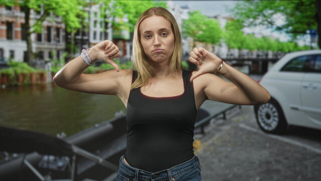 Woman wearing jeans and tanktop showing thumbs down gesture and pouting on a street canal beside a parked car; disapproval rejection attitude.