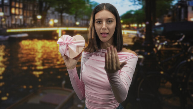 Young woman holding heart shaped gift and beckoning with open hand on street bridge at night; romantic invitation.