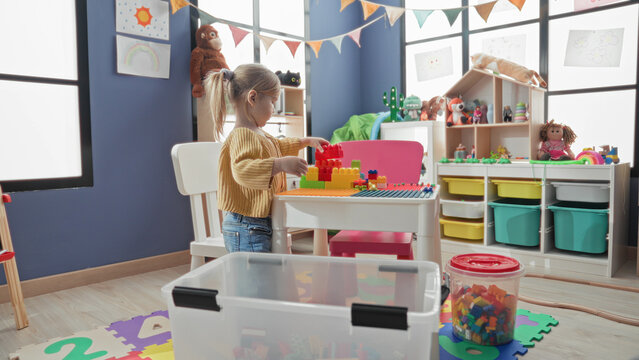 Girl toddler arranges colorful plastic blocks on a table with bare hands in a home playroom inside building; concentration play creativity.