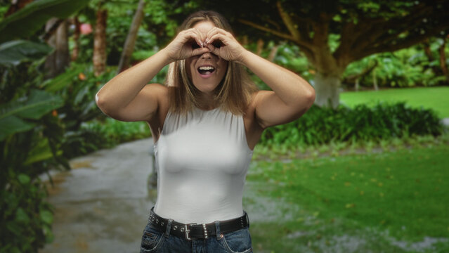 Young blonde woman forming binoculars with hands to eyes, white tank top and jeans on a path under trees in a forest; playful curiosity.