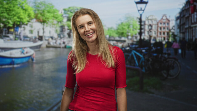 Woman smiling at camera, standing by a canal with boat and bicycle on a street lined with lampposts and buildings; cheerful warmth.