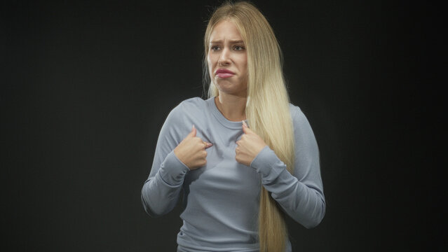 Young blonde woman points both thumbs to her chest with puzzled grimace and pursed lips wearing light blue sweater against black backdrop studio; confusion doubt.
