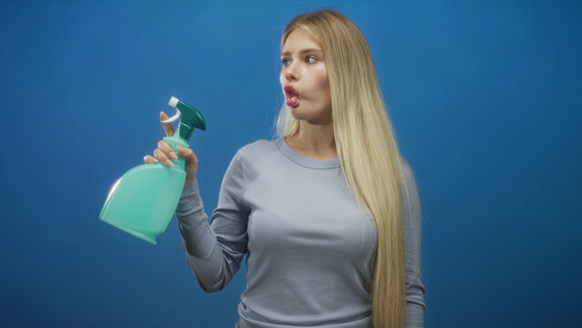 Woman holding spray bottle with puckered lips and hand gripping nozzle in plain blue studio, long blonde hair visible; playfulness freshness.