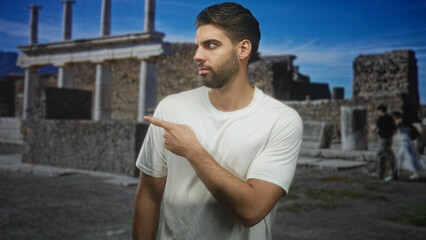 Fototapeta premium Man with beard points finger to ancient building ruins while wearing a white tshirt, side profile visible; curiosity discovery.